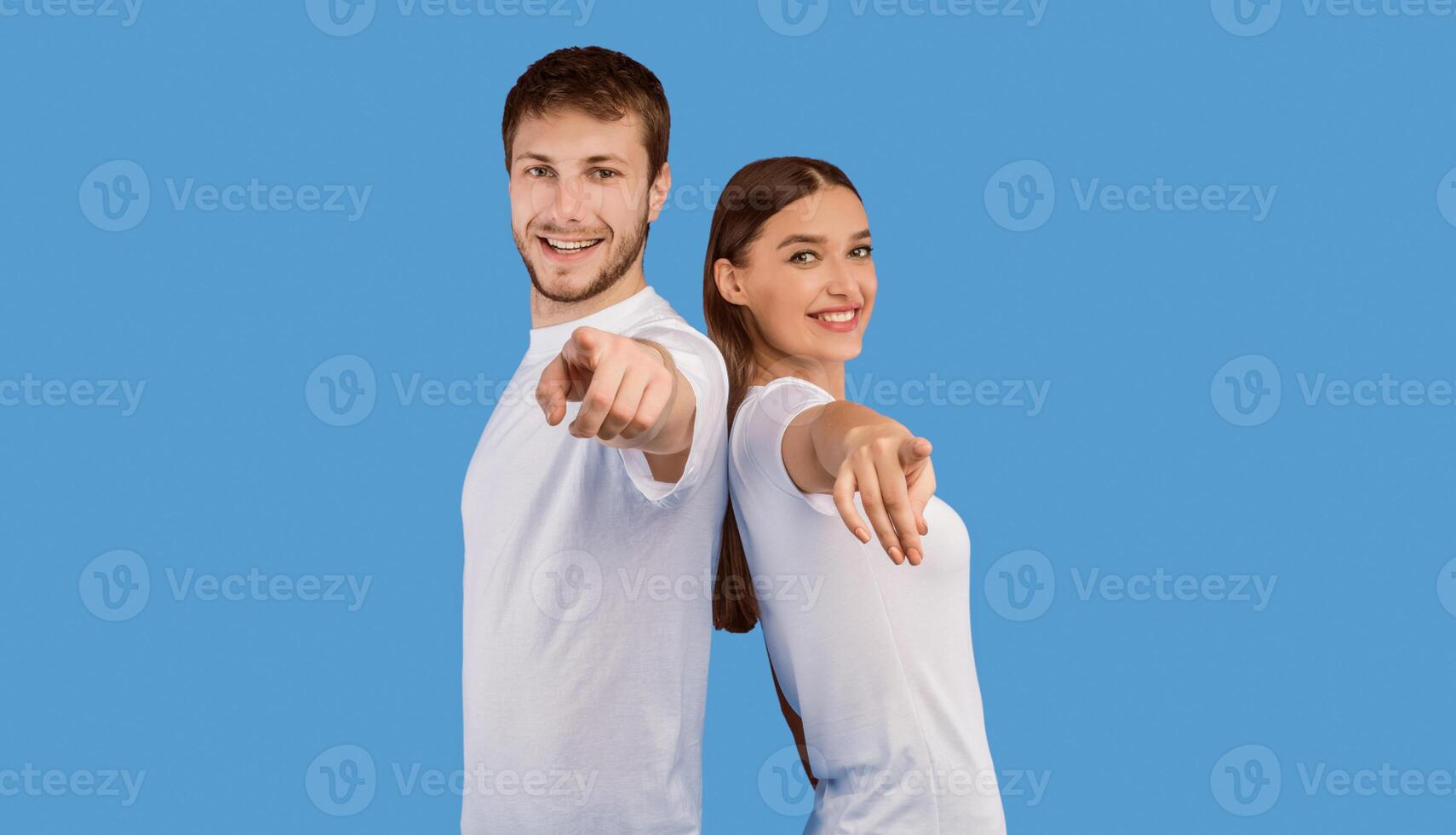 Hey, You. Portrait of cheerful casual friends pointing finger at camera, standing back to back on purple wall, copyspace photo