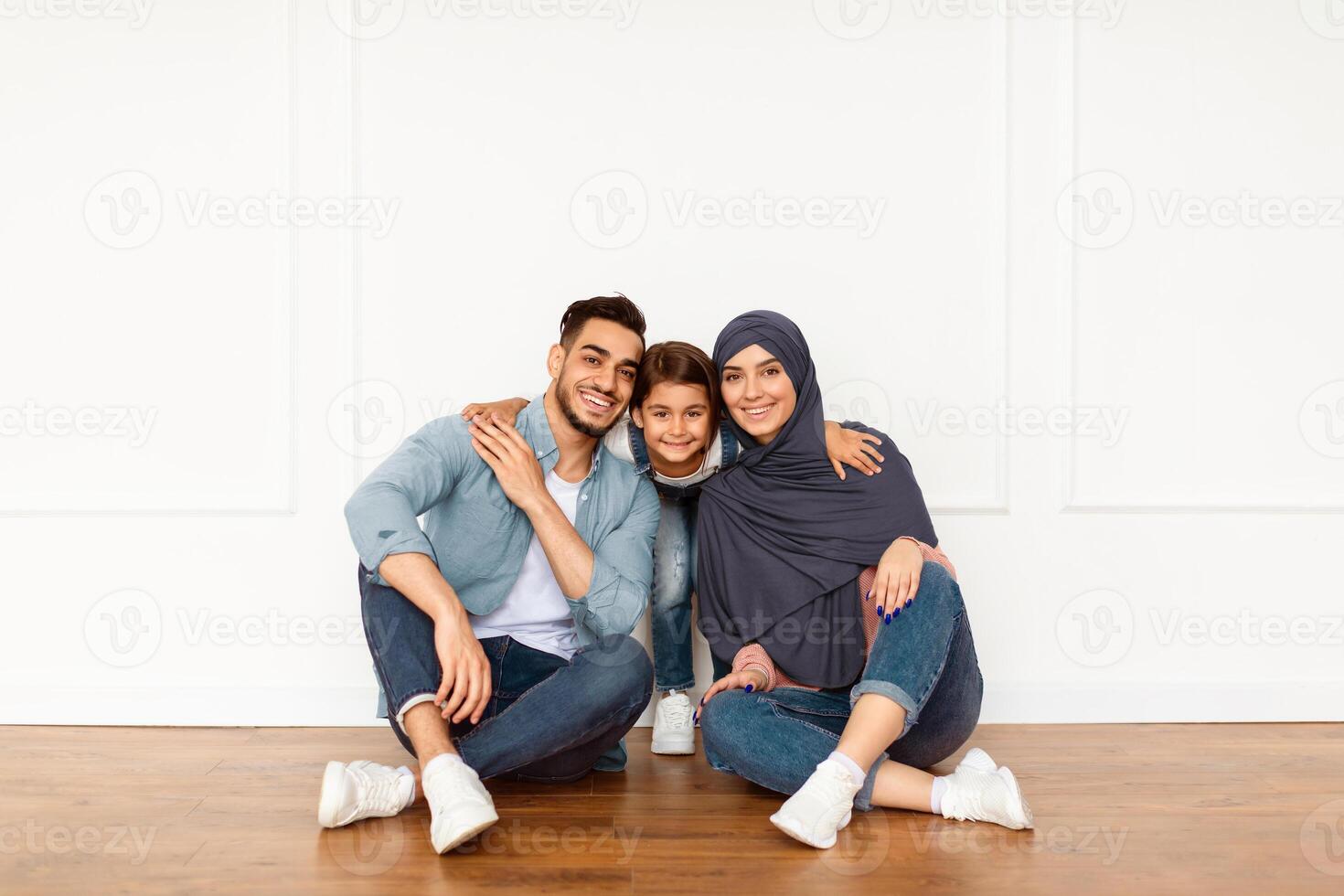 Portrait of loving happy Middle Eastern family with little preschooler kid sitting near white wall looking at camera and smiling, parents enjoy time with small child posing together in modern new home photo