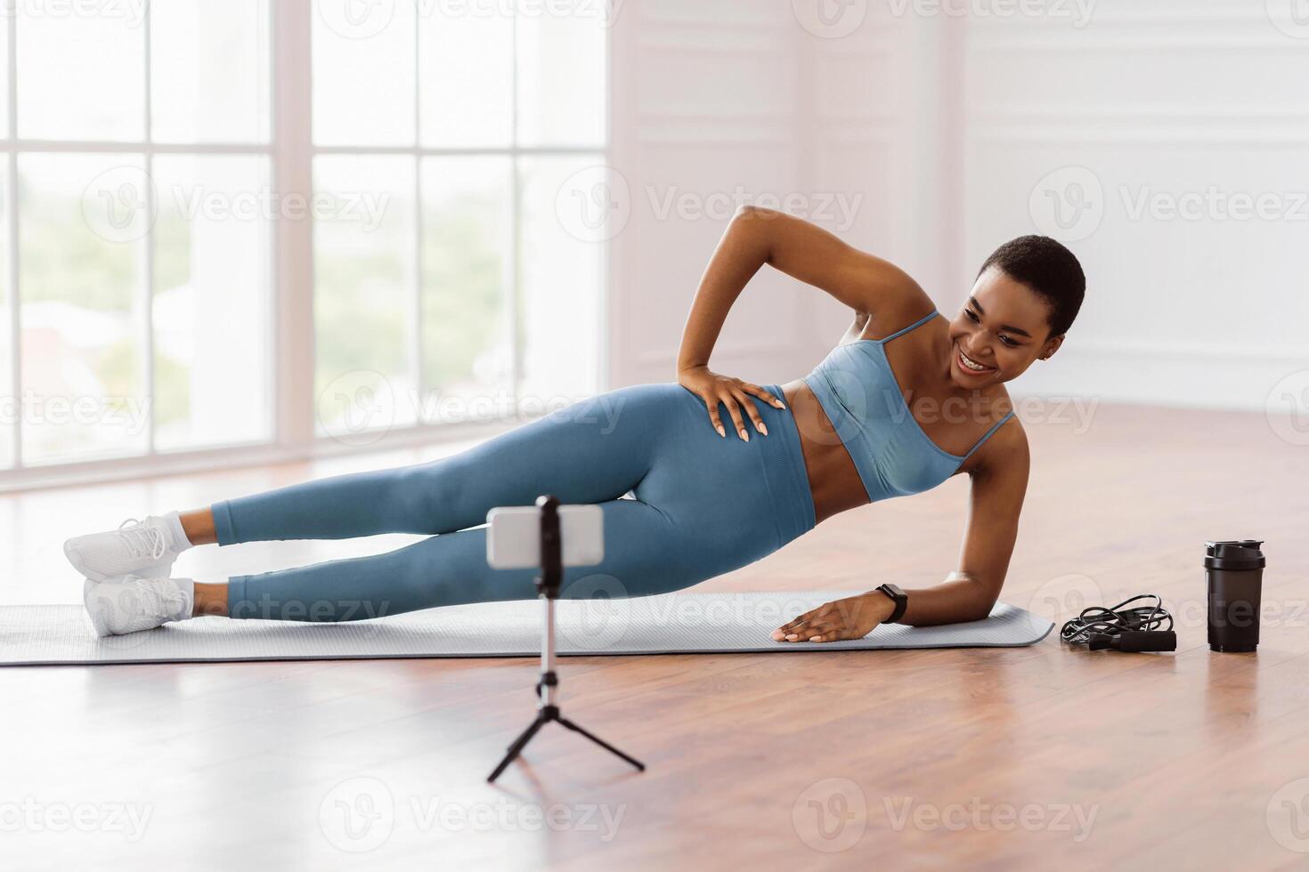 Online Workout. Smiling African Lady Standing In Side Plank Position On Yoga Mat Near Window, Looking At Mobile Phone On Tripod, Watching Tutorial Or Recording For Blog, Having Chat With Coach photo