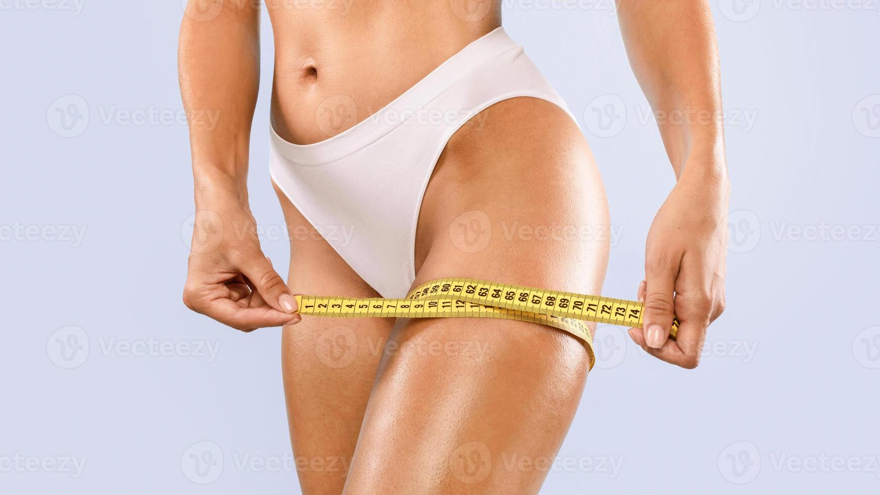 Time For Slimming Diet And Weight Loss Concept. Closeup cropped view of unrecognizable fit woman in white panties using measure tape measuring her thigh isolated on blue studio background, banner photo
