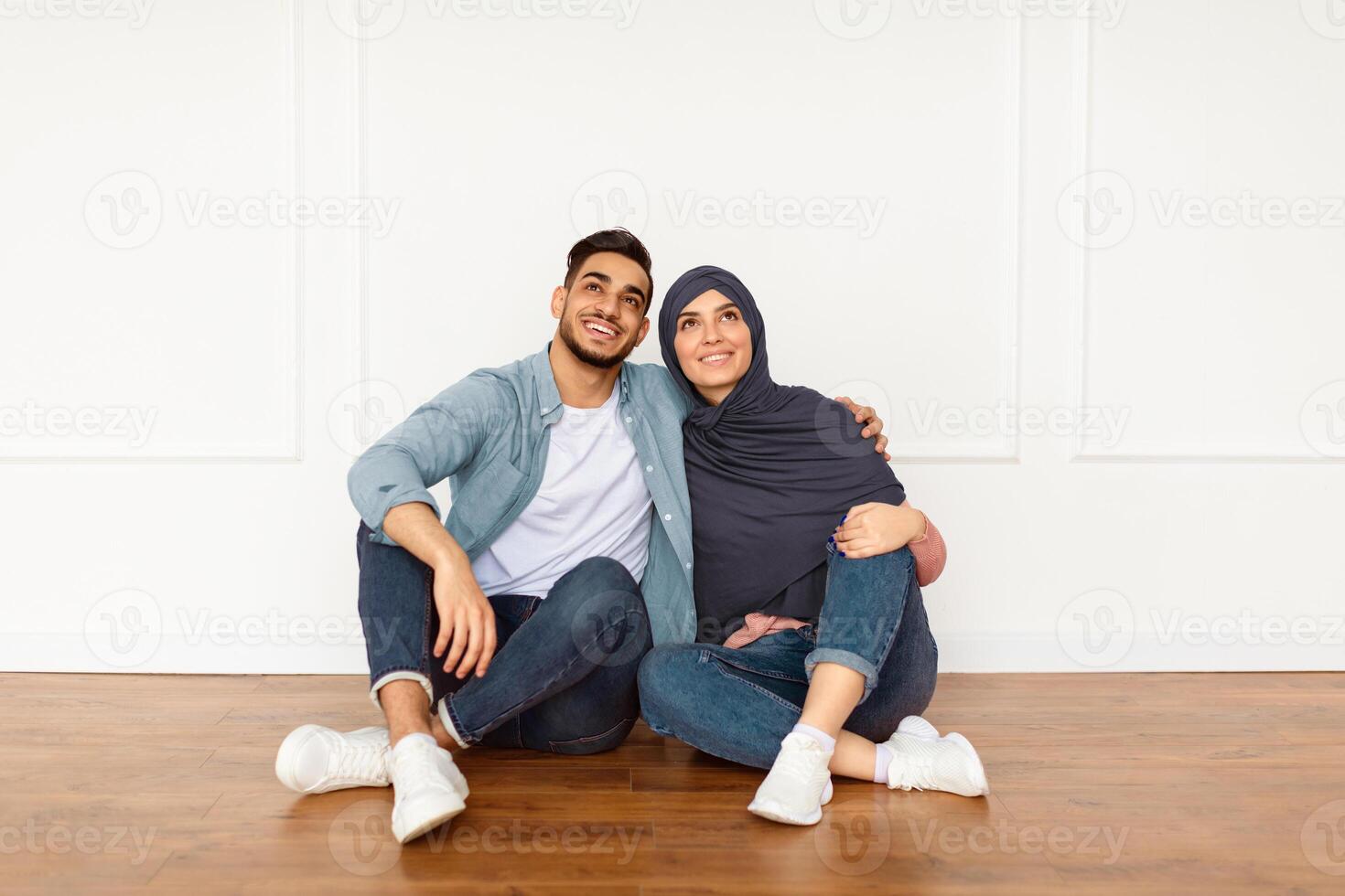 Dreaming About Future. Portrait of smiling muslim couple sitting on floor leaning on white wall in new apartment, looking up at free copy space. Cheerful guy and lady in hijab planning repair photo