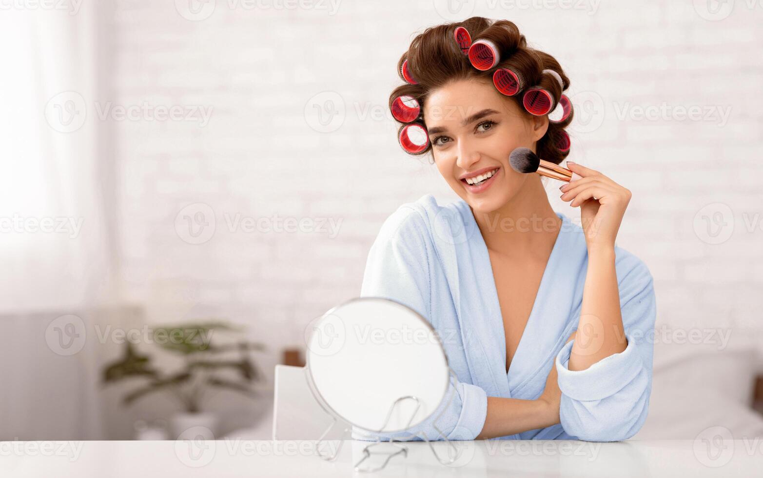 Attractive Lady With Hair Curlers Making Makeup Using Cosmetic Brush Applying Powder Sitting In Bedroom At Home, Smiling To Camera. Female Daily Make Up And Beauty Routine Concept photo