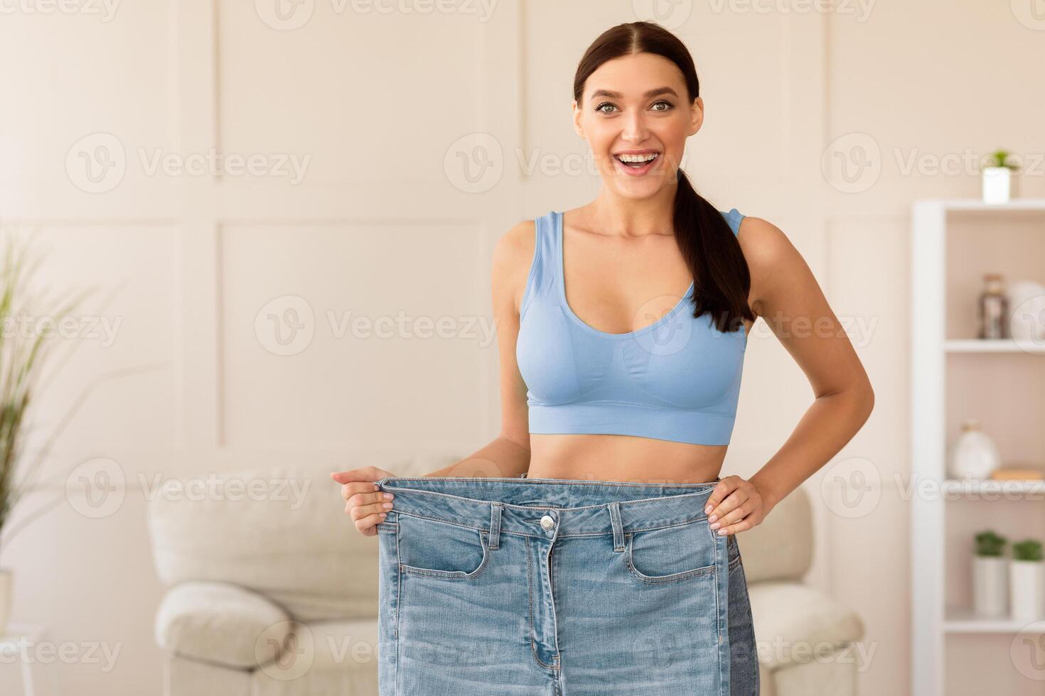Skinny Woman Comparing Old Oversize Jeans After Dieting And Successful Weight Loss Standing At Home. Free Space photo