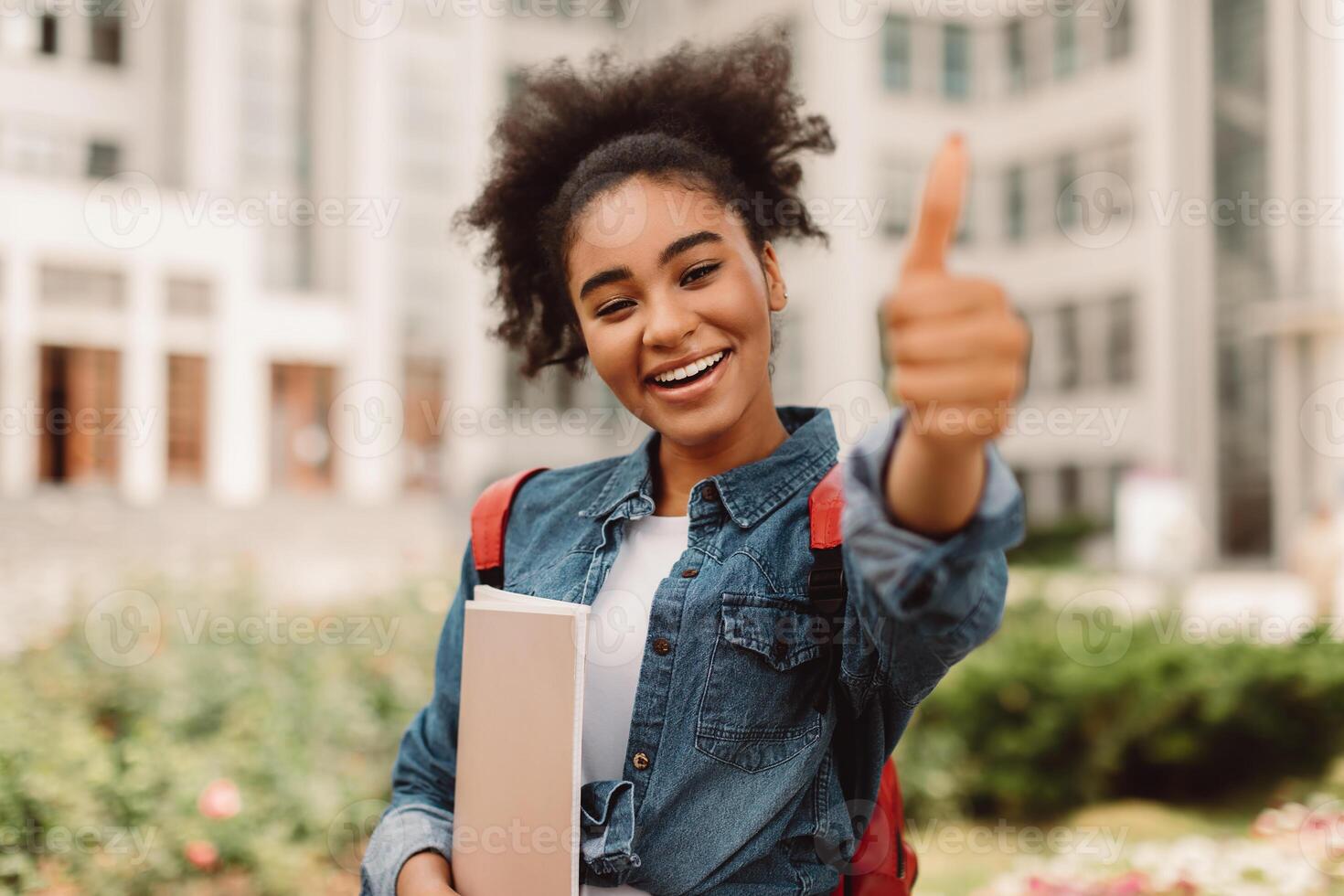 Happy Black Female Student Gesturing Thumbs-Up Holding Books Posing With Backpack On University Campus Territory Outside. I Like My College, Education Concept. Selective Focus photo