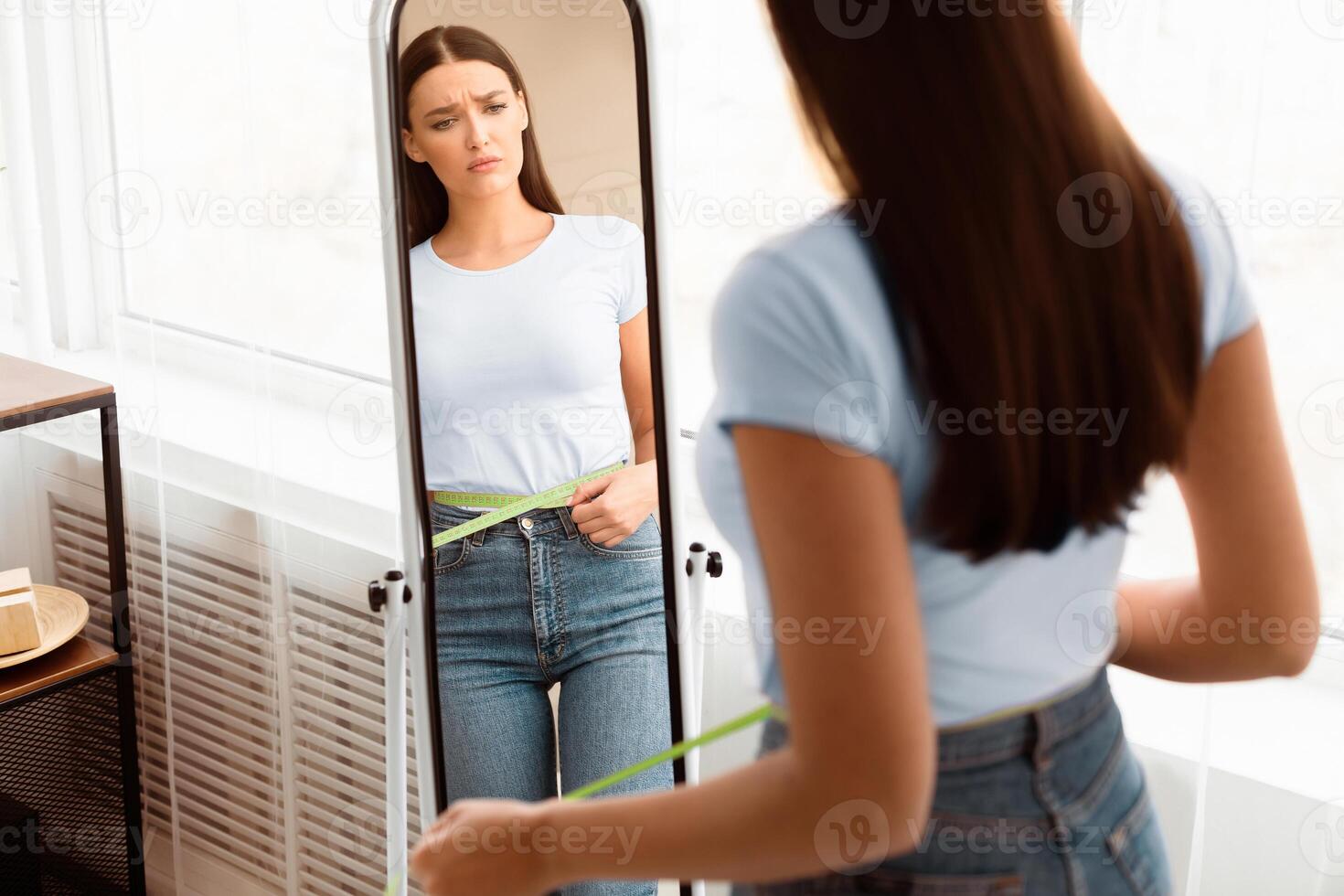 Sad Woman Measuring Waist With Tape After Weight Gain Standing In Front Of Mirror At Home. Slimming Concept. Free Space photo