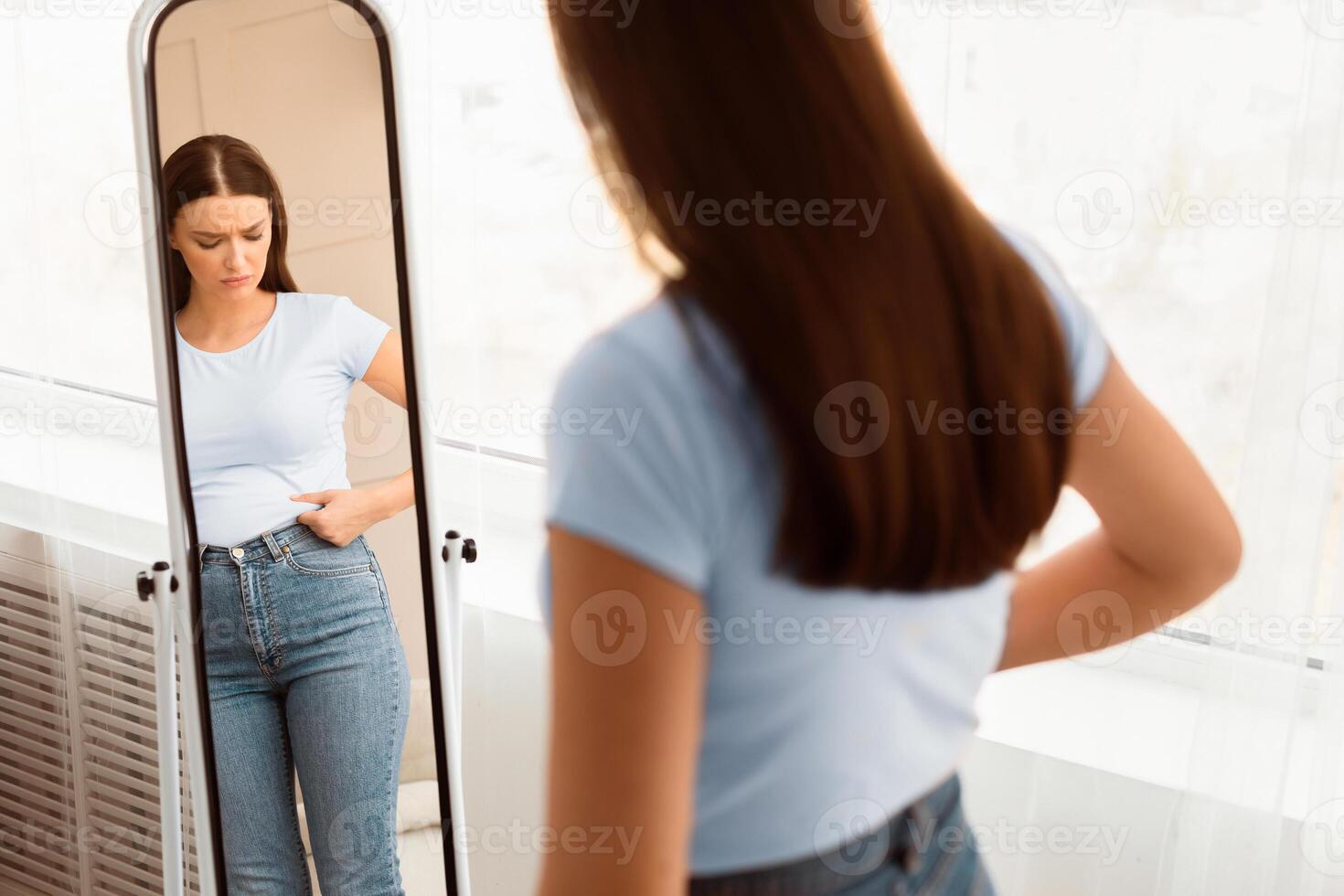 Unhappy Woman Gaining Excess Weight Touching Drooping Belly Standing At Mirror At Home. Unsuccessful Diet. Selective Focus photo