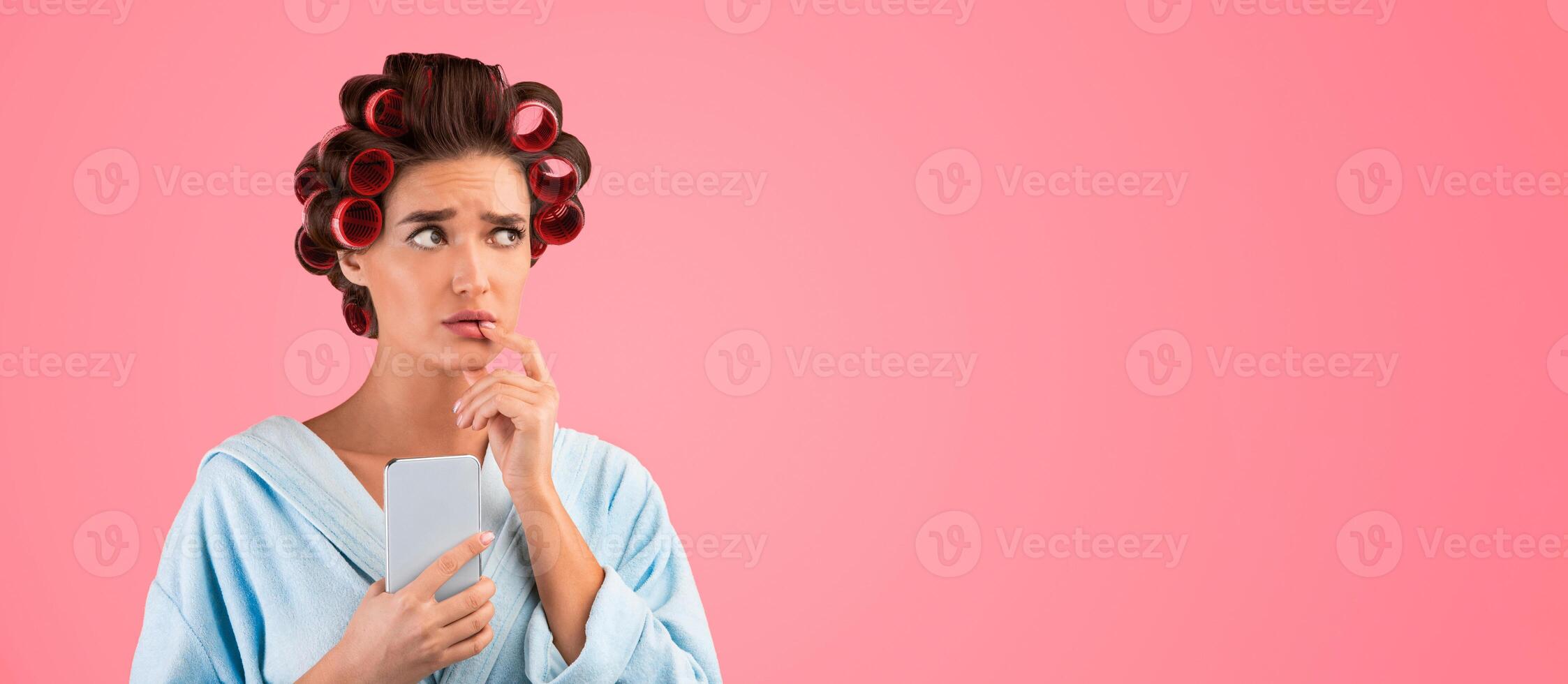 Puzzled Housewife Holding Smartphone Thinking Standing On Yellow Studio Background. Pensive Lady With Hair Curlers Using Cellphone And Mobile Phone Application. Panorama, Empty Space For Text photo
