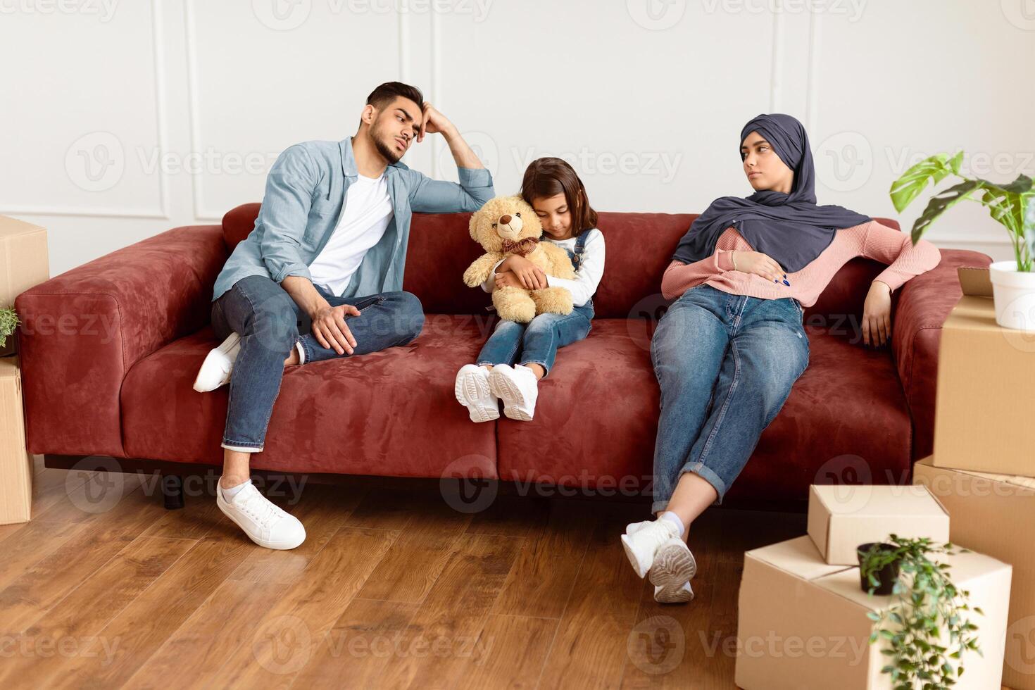 Sad family of three people sitting on sofa with cardboard boxes around, being tired on moving day. Exhausted husband, wife and daughter kid taking break from unpacking things. Renting issues concept photo