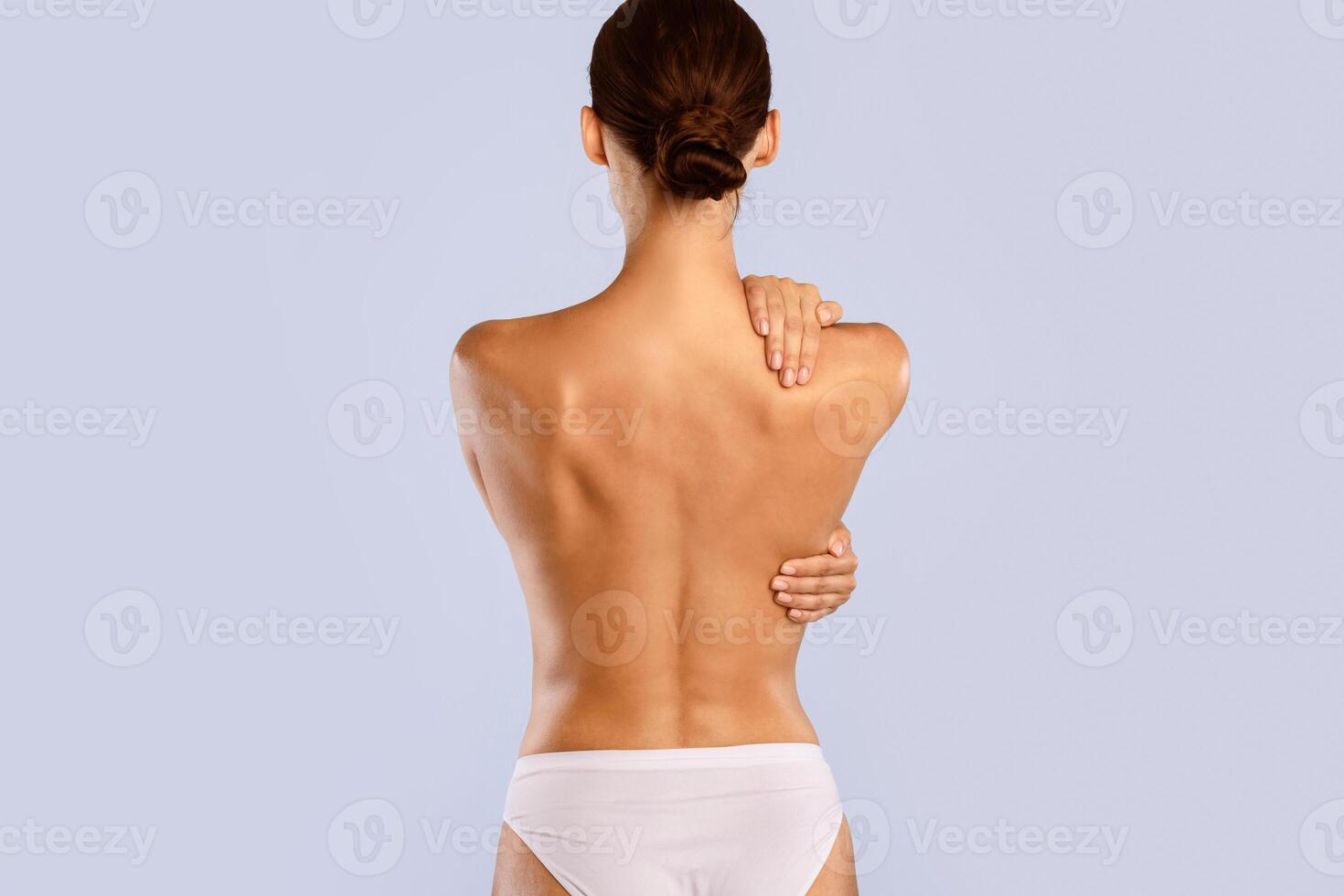 Rear back view of young naked woman in white panties embracing herself, touching shoulder and spine, massaging muscles, standing isolated over blue studio background. Beauty Treatment photo