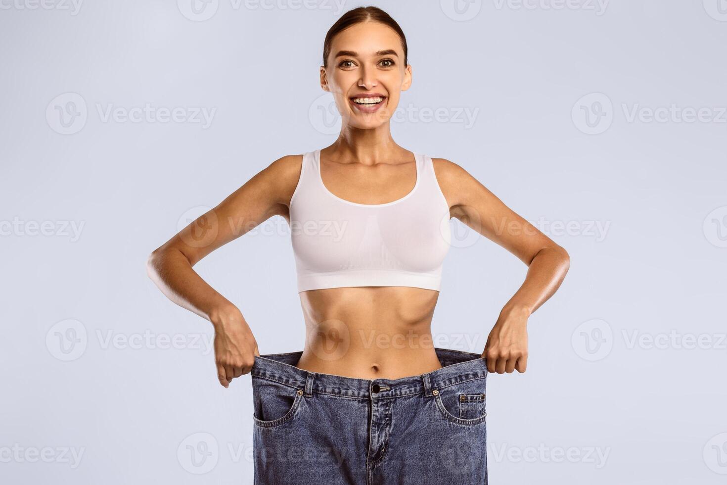 Slimming Concept. Portrait Of Excited Young Woman Wearing Too Big Denim Jeans After Successful Weight Loss, Posing and Looking At Camera Standing Isolated Over Blue Studio Background, Free Space photo
