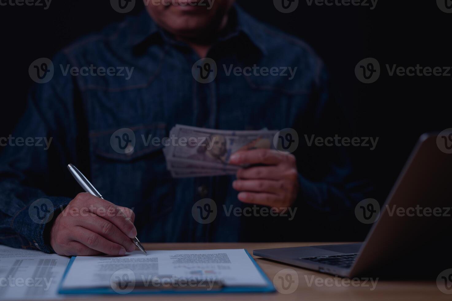 A man is sitting at a desk with a laptop and a stack of cash. He is writing something on a piece of paper photo