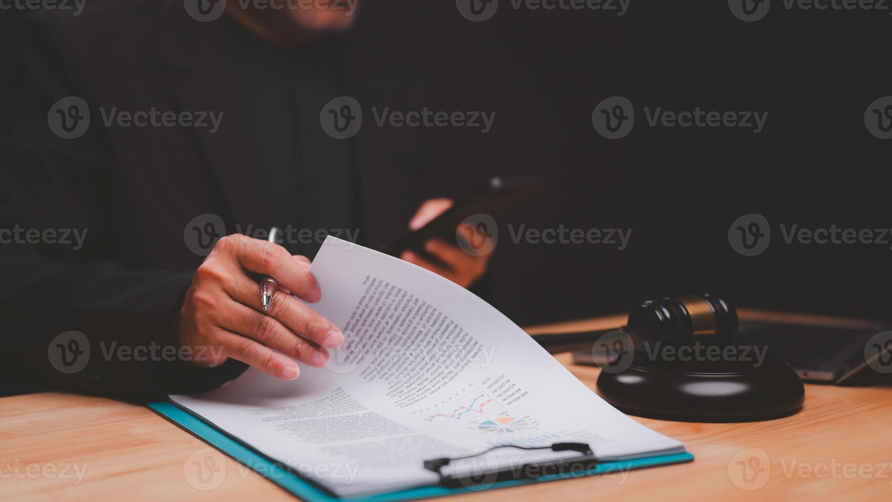 Legal professional examines important documents while holding smartphone, surrounded by gavel and laptop, symbolizing intersection of law and technology in modern practice photo