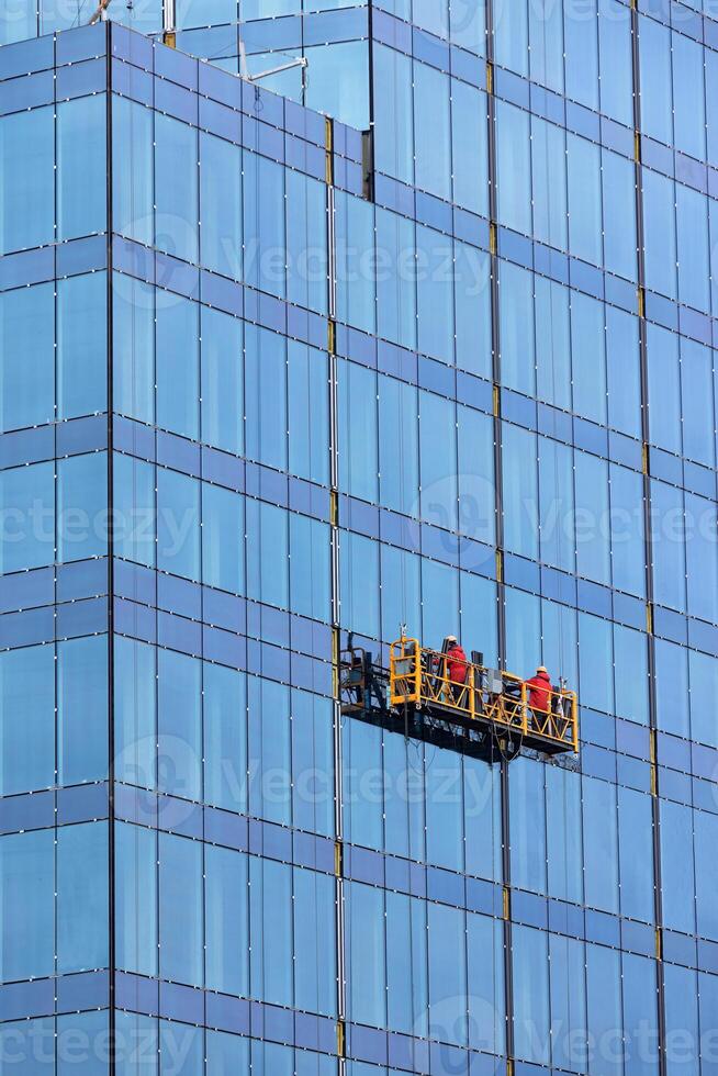 Builders in red overalls are mounting the glass facade of a multi-storey building on a suspended cradle. photo