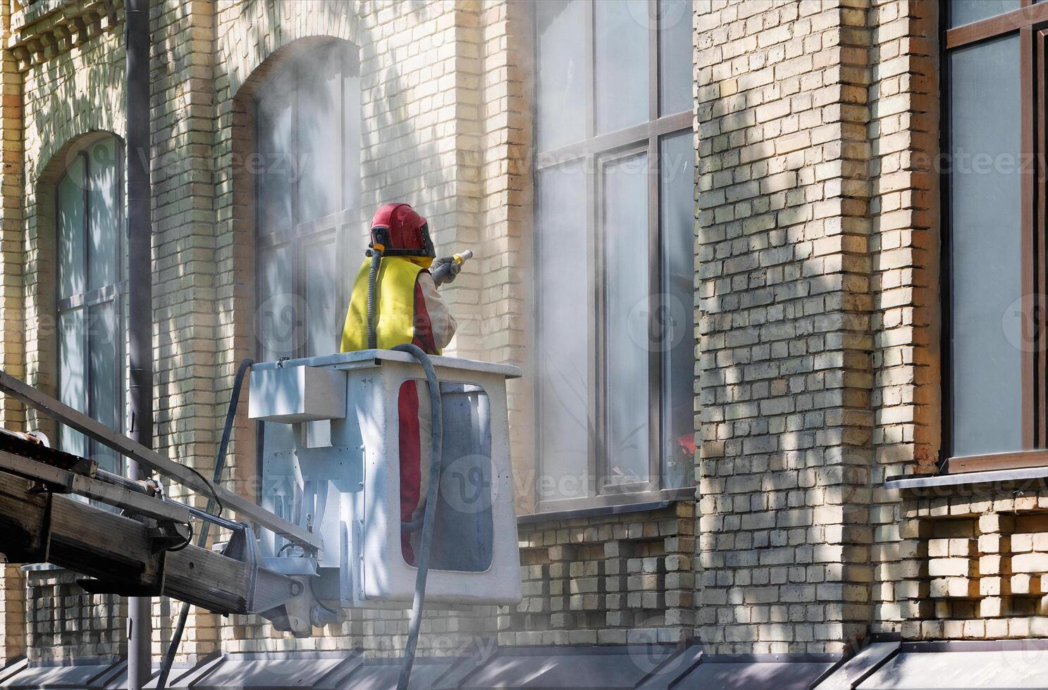 A worker in a protective suit cleans the facade of an old brick house with an industrial sandblaster. photo
