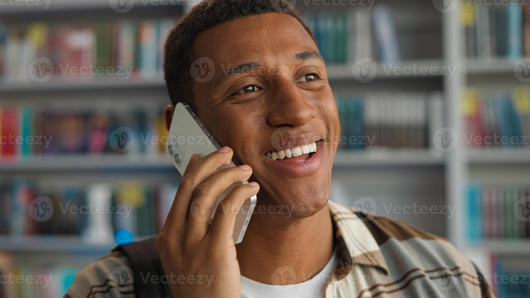 Happy African American man talking mobile phone smartphone technology communicate conversation library student learner university chatting smiling male guy bookshelf educational study calling laughter photo