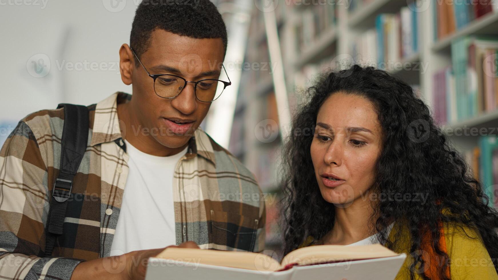 Two students African American man Arabian Latino woman together reading book textbook library talking discussion university college classmates speak study learning hobby girl guy high school bookstore photo