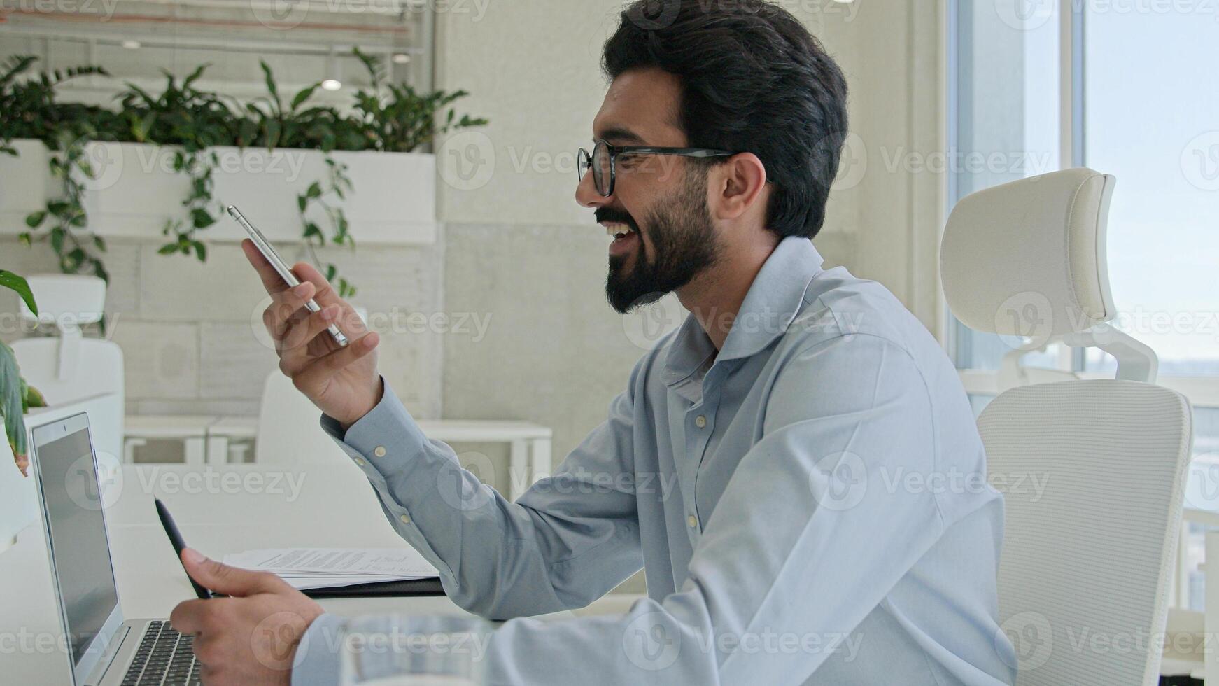 Busy multitasking Arabian businessman manager consultant Indian muslim man talk mobile phone check information on laptop computer work consulting client smartphone call business consultation in office photo