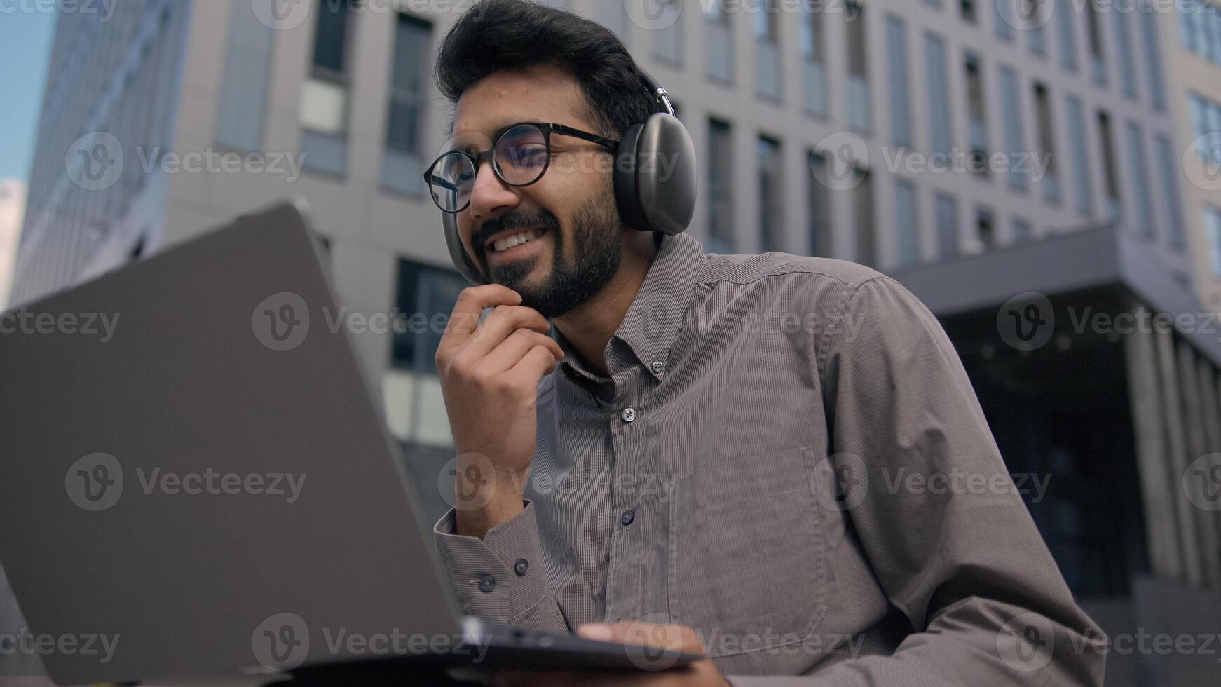 Arabian Indian man listen music educational webinar in headphones outdoors male businessman freelancer work with laptop develop business project online studying smiling guy student e-learning in city photo