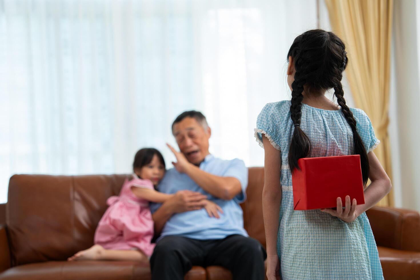 Little girl hiding present box behind back while making surprise elderly grandparents, Cheerful elderly man smiling and gesticulating while receives a surprise gift from granddaughter for celebrations photo