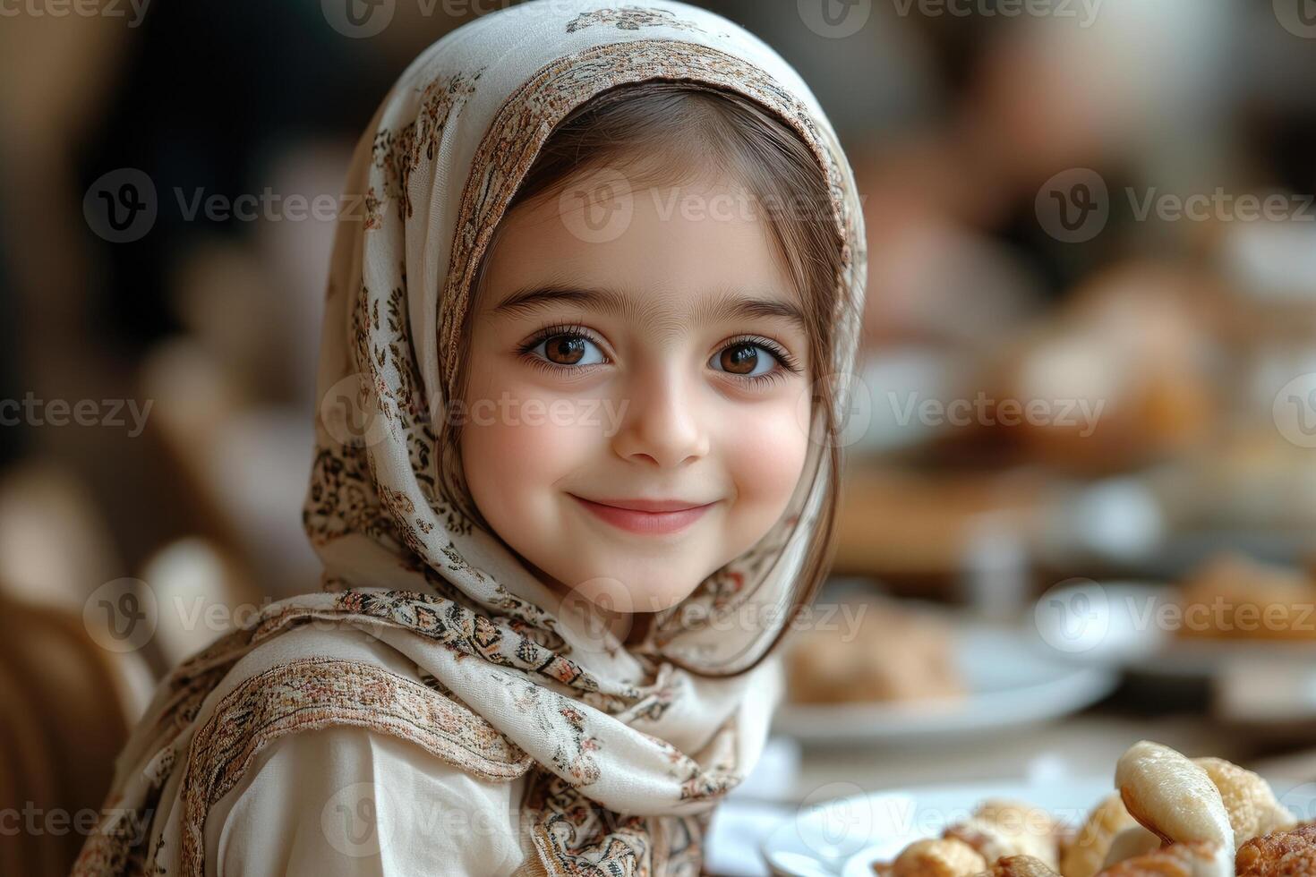 A little girl wearing a headscarf sitting at a table with food photo
