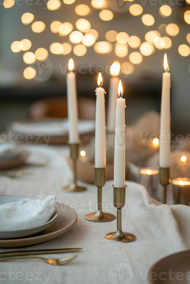 Tall white candles in simple brass holders illuminate a beautifully arranged Easter table. Muted ceramic plates and soft linen runners create a cozy atmosphere with glowing lights above photo