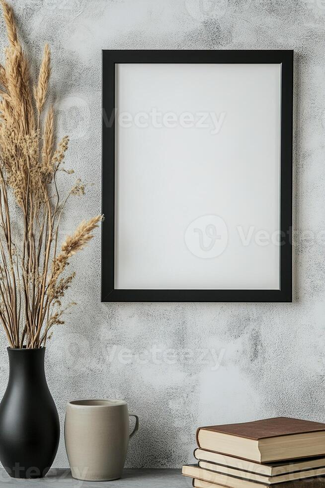 A sleek picture frame with a thin black border is mounted on a textured concrete wall. Nearby, a ceramic vase holds dried grass, and a stack of books adds a touch of minimalistic style photo