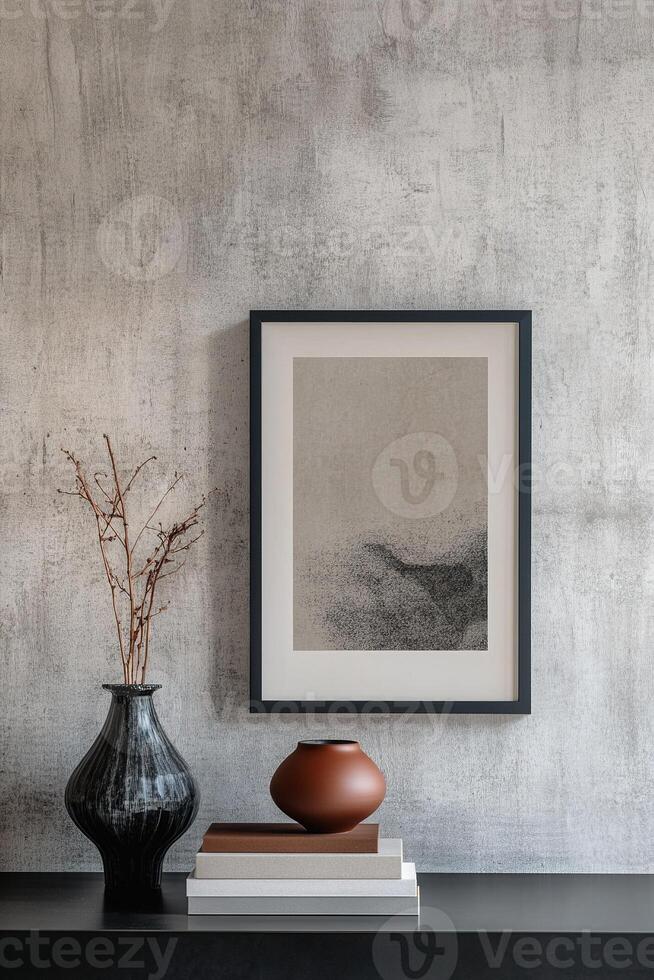 A sleek picture frame with a thin black border hangs against a textured concrete wall. It is accompanied by a ceramic vase and a neatly stacked set of books, creating a chic minimalist aesthetic photo