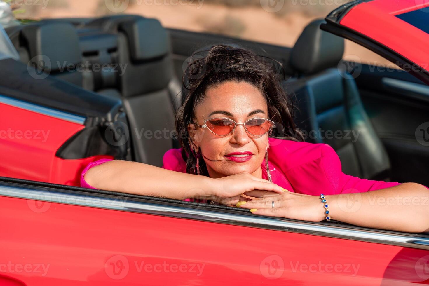A woman in a pink jacket is sitting in a red convertible. She is wearing sunglasses and has her hand on her hip. Scene is relaxed and carefree. photo