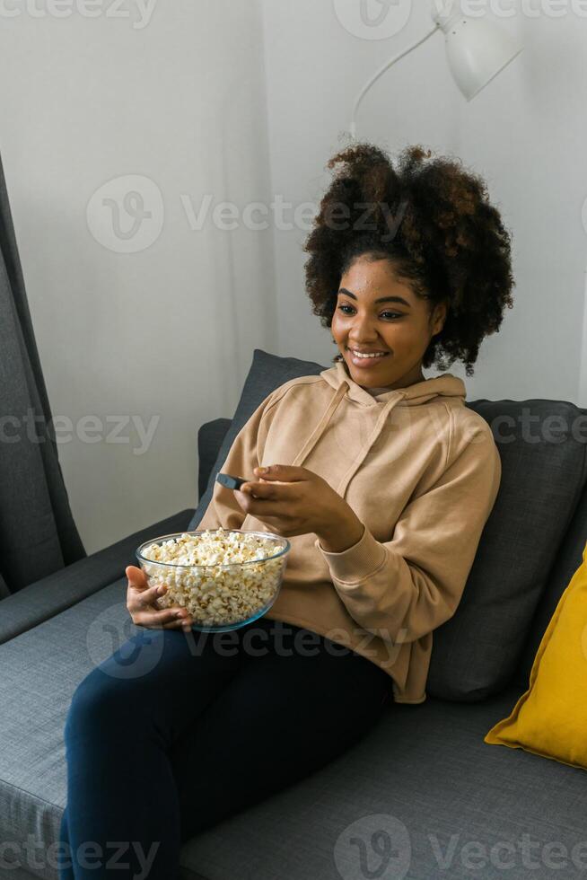 Young african american woman with a remote control in one hand and popcorn watches a television program. Cheerful enjoyment of leisure time. Streaming service, film and tv show concept. photo