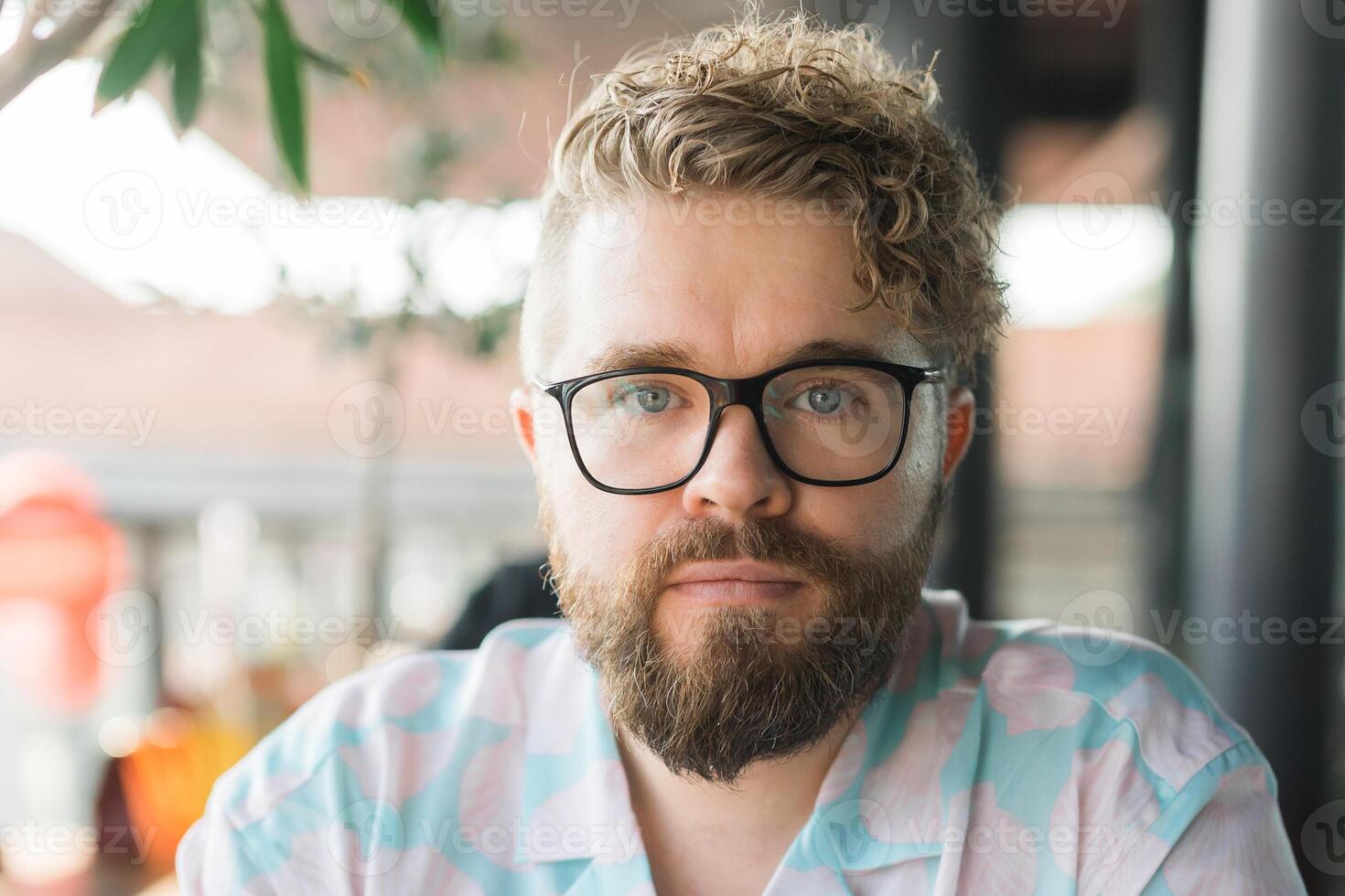 Close-up portrait of a man with glasses, curly hair, and beard, sitting outdoors at a cafe. Neutral expression and natural lighting highlight individuality and a calm, thoughtful demeanor. Millennial photo