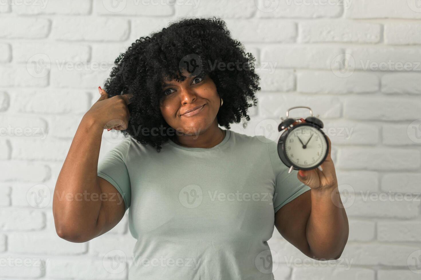 Surprised african american woman holding alarm clock amazed with open mouth and surprise happy face at home, on brick background. Being late and time passing quickly concept photo