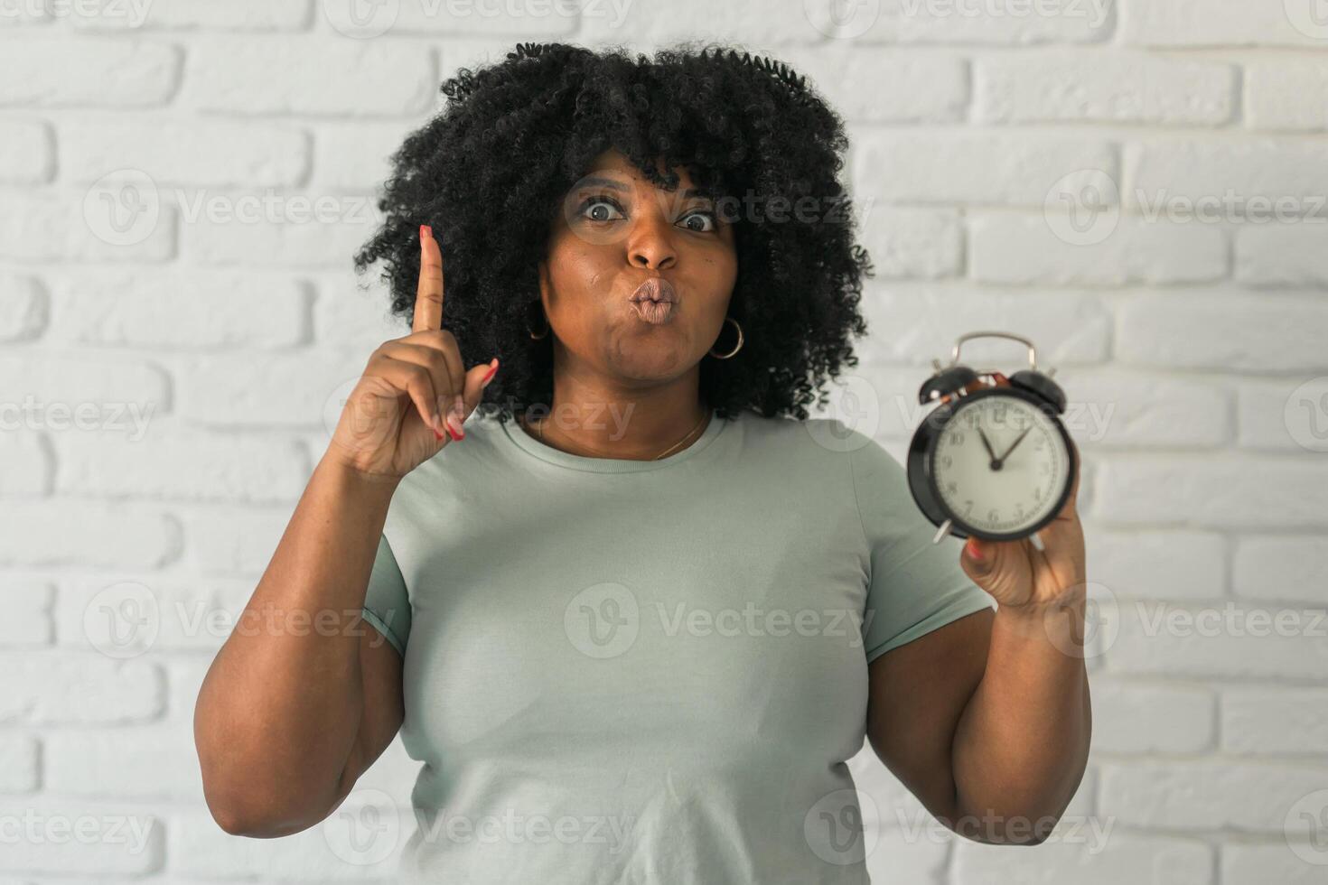 Surprised african american woman holding alarm clock amazed with open mouth and surprise happy face at home, on brick background. Being late and time passing quickly concept photo