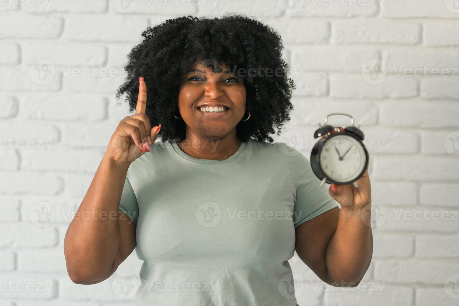 Surprised african american woman holding alarm clock amazed with open mouth and surprise happy face at home, on brick background. Being late and time passing quickly concept photo