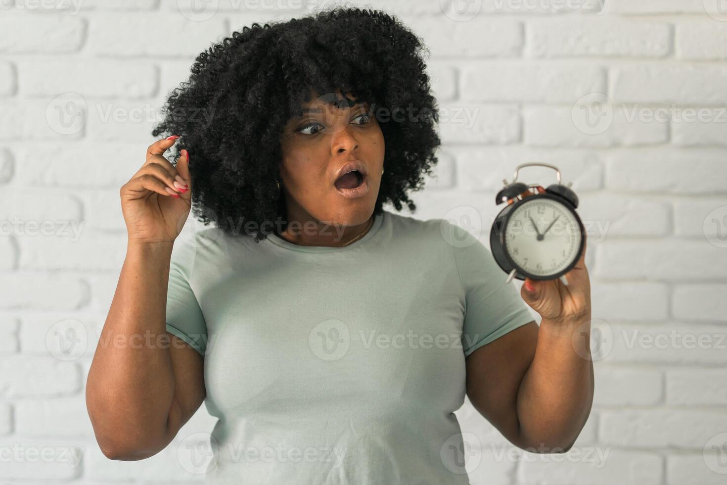 Surprised african american woman holding alarm clock amazed with open mouth and surprise happy face at home, on brick background. Being late and time passing quickly concept photo