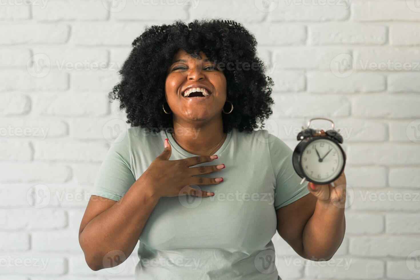 Surprised african american woman holding alarm clock amazed with open mouth and surprise happy face at home, on brick background. Being late and time passing quickly concept photo