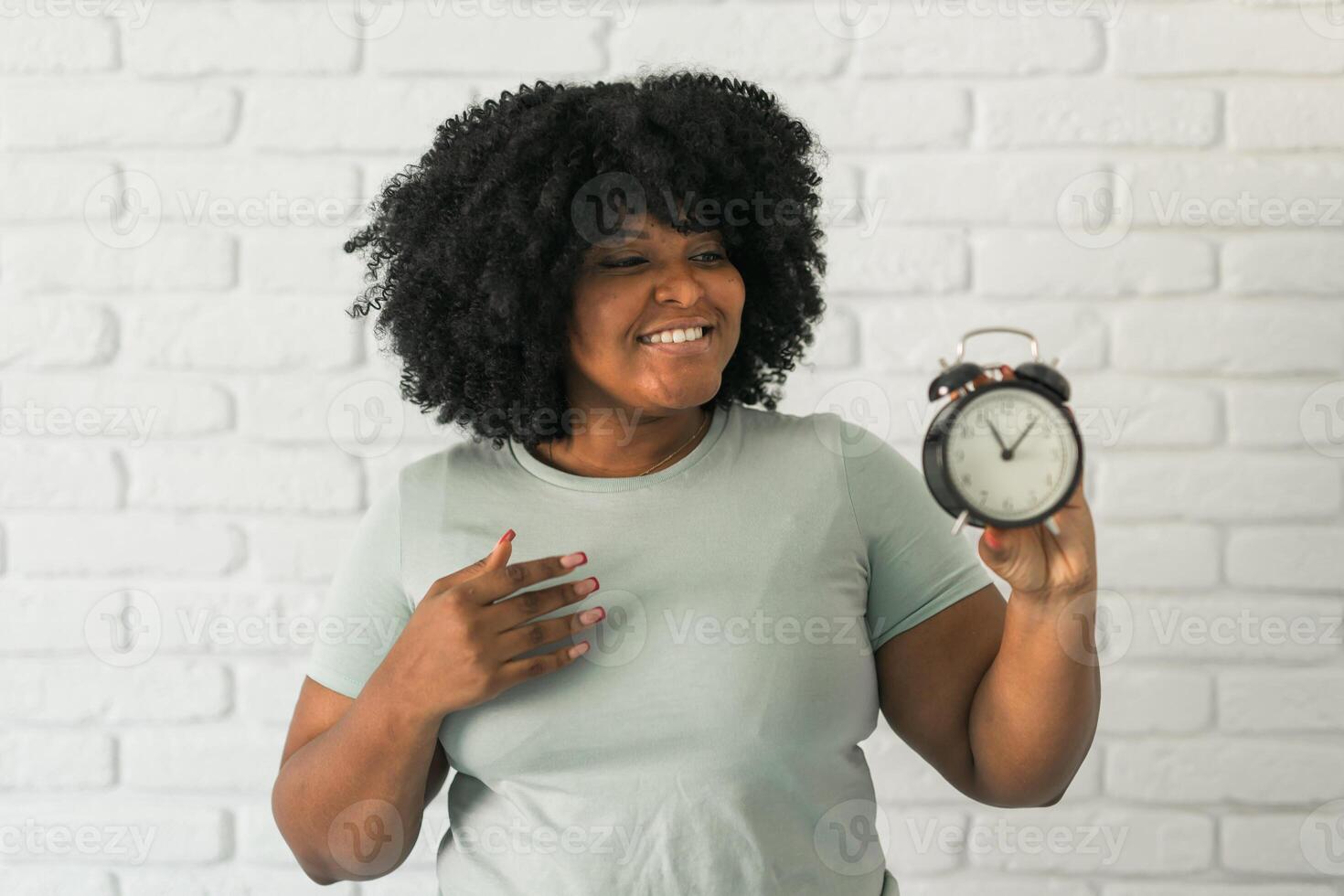 Surprised african american woman holding alarm clock amazed with open mouth and surprise happy face at home, on brick background. Being late and time passing quickly concept photo
