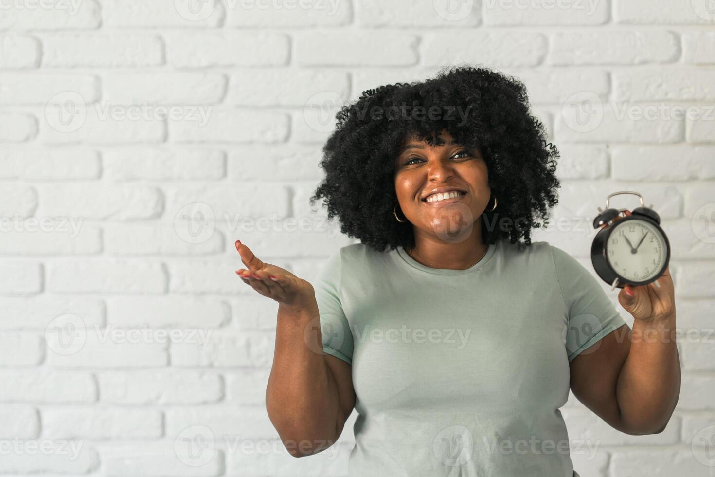 Surprised african american woman holding alarm clock amazed with open mouth and surprise happy face at home, on brick background copy space. Being late and time passing quickly concept photo