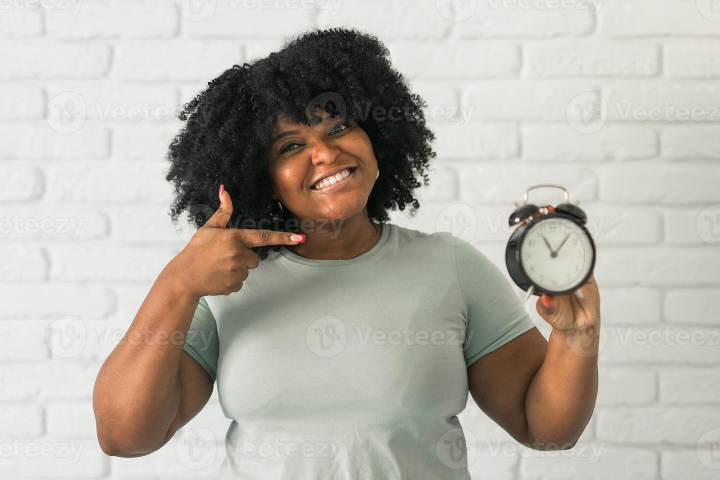 Surprised african american woman holding alarm clock amazed with open mouth and surprise happy face at home, on brick background. Being late and time passing quickly concept photo