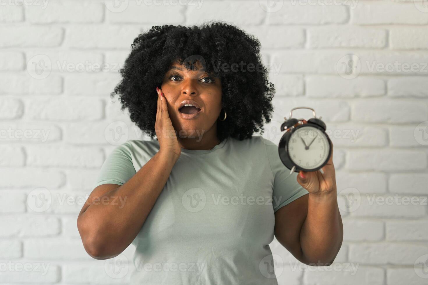 Surprised african american woman holding alarm clock amazed with open mouth and surprise happy face at home, on brick background. Being late and time passing quickly concept photo