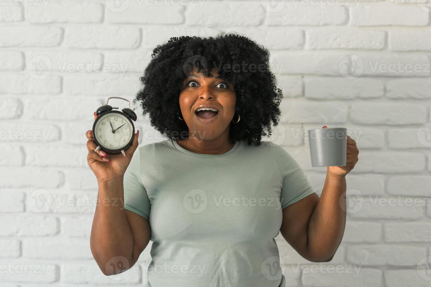 Surprised african american woman holding alarm clock amazed with open mouth and surprise happy face at home, on brick background. Being late and morning time and time passing quickly concept photo