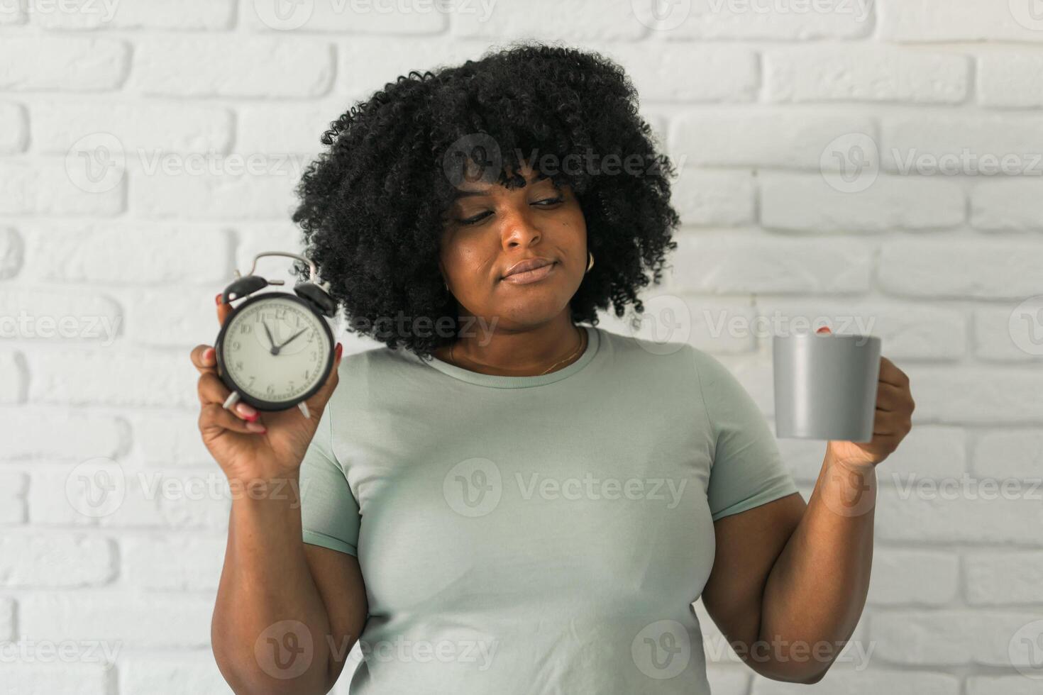 Happy african american woman holding alarm clock amazed with open mouth and surprise happy face at home, on brick background. Being late and morning time and time passing quickly concept photo