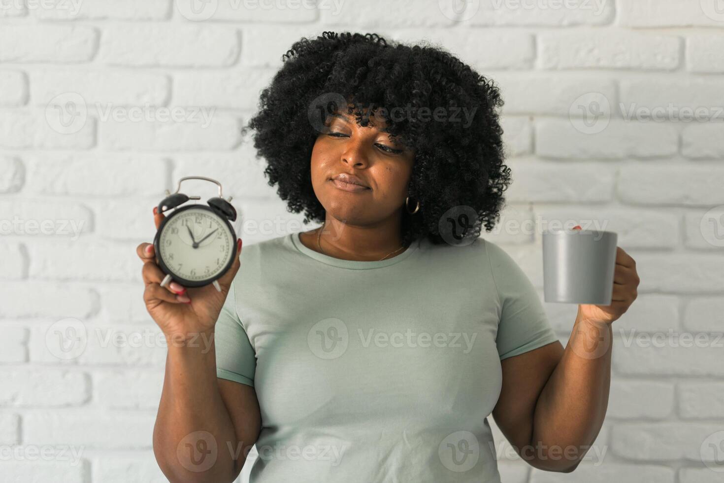Happy african american woman holding alarm clock amazed with open mouth and surprise happy face at home, on brick background. Being late and morning time and time passing quickly concept photo