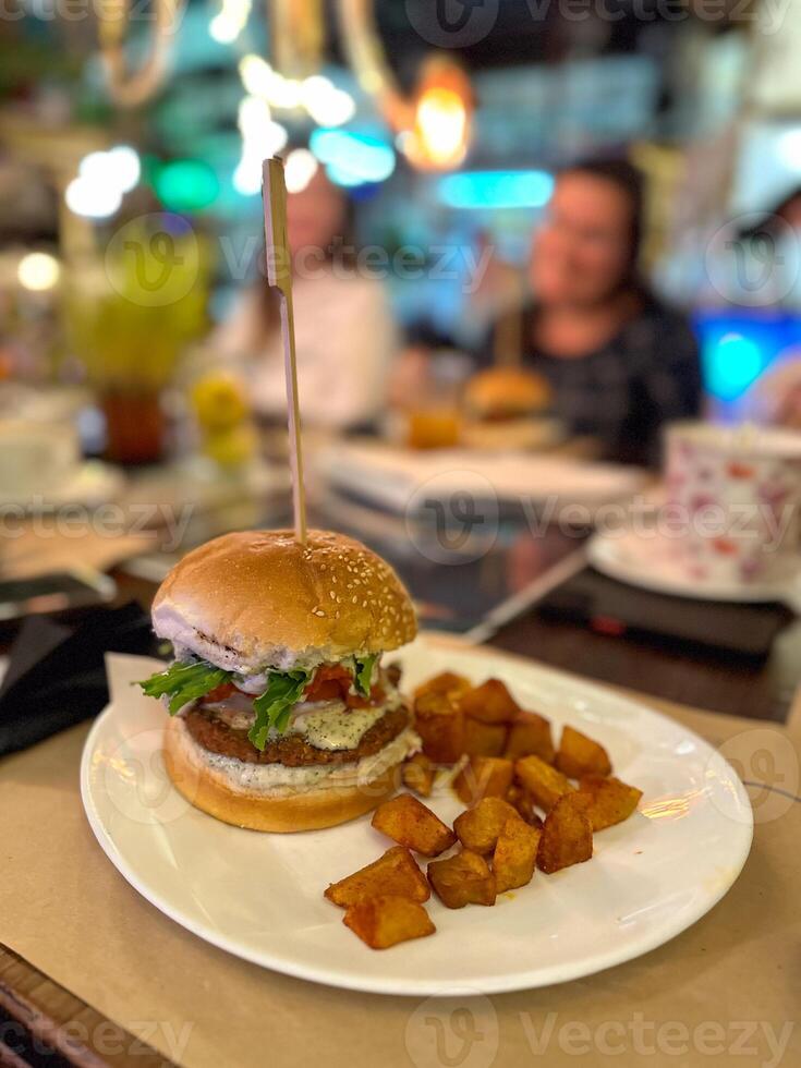 Juicy vegan burger with fresh toppings is plated alongside crispy golden potato cubes. Alternative plant-based meat in burger photo