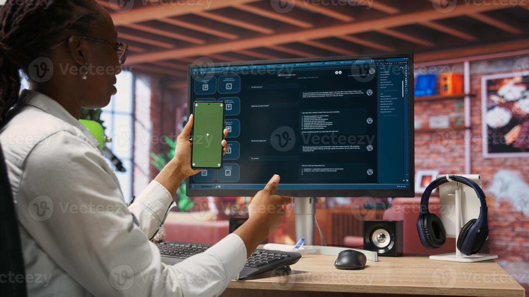 Woman seated at desk using AI LLM chatbot software and green screen phone in modern urban home office. Person uses large language model program on computer and mockup smartphone in apartment, camera B photo
