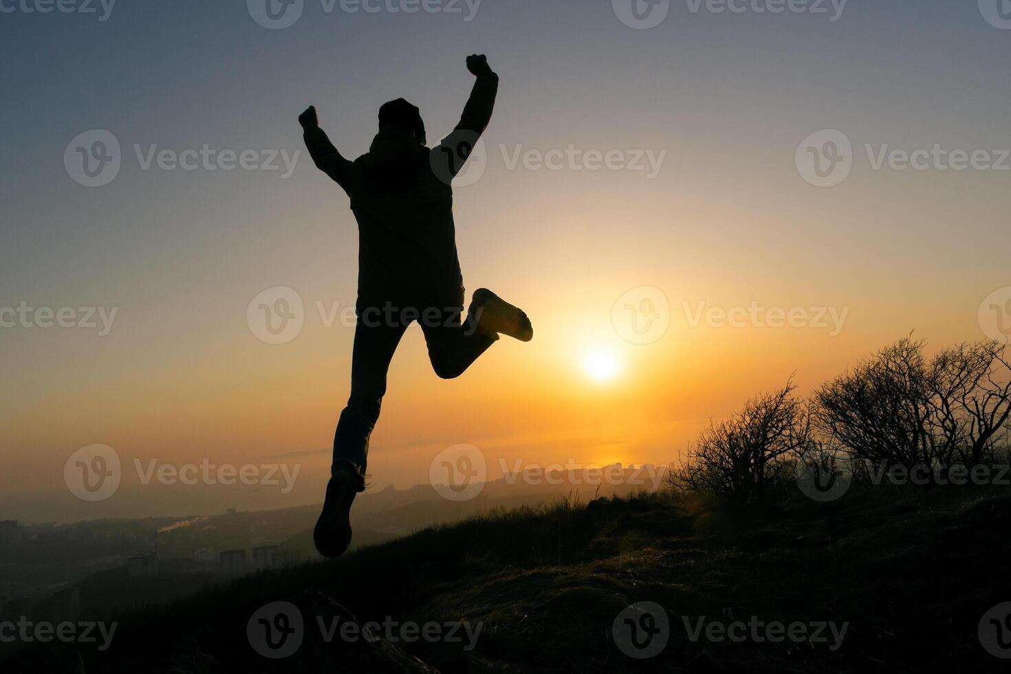 Girl jumping in a jacket and a hat on the top of the mountain during sunset. Silhouette in the rays of the sun photo