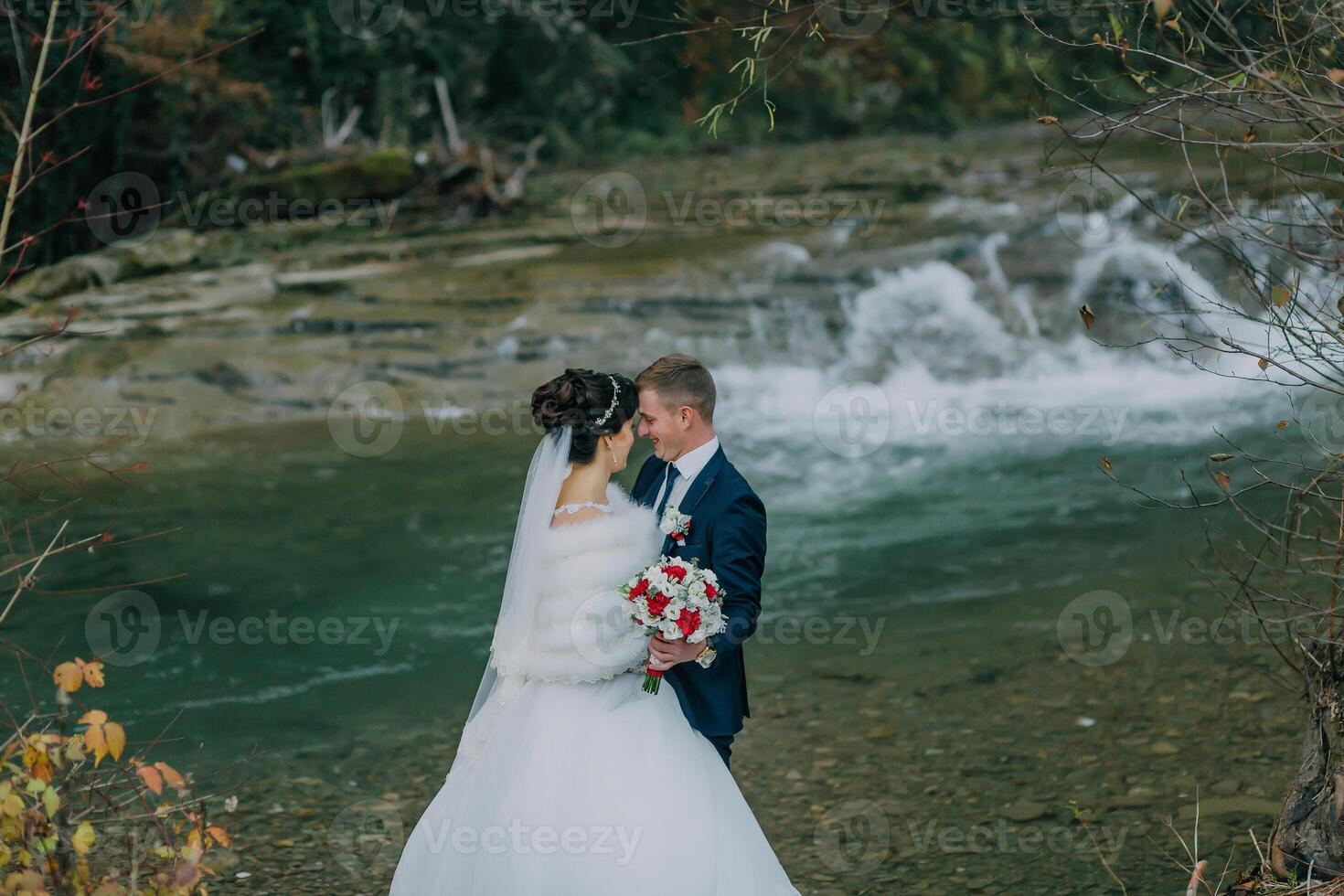 A bride and groom are standing in front of a waterfall, holding hands photo