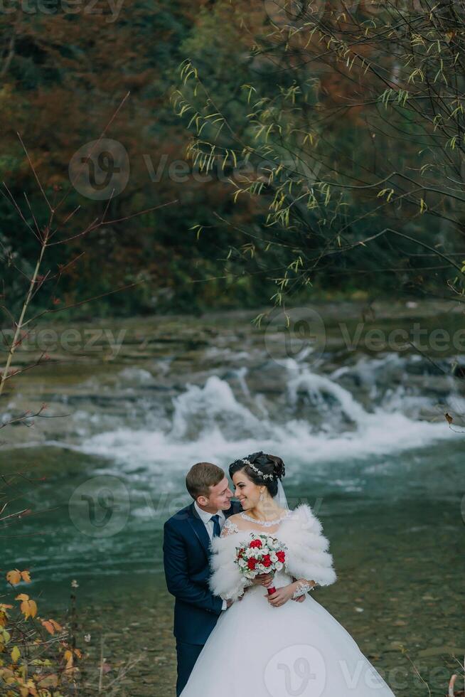 A bride and groom are standing by a river, holding hands and smiling photo