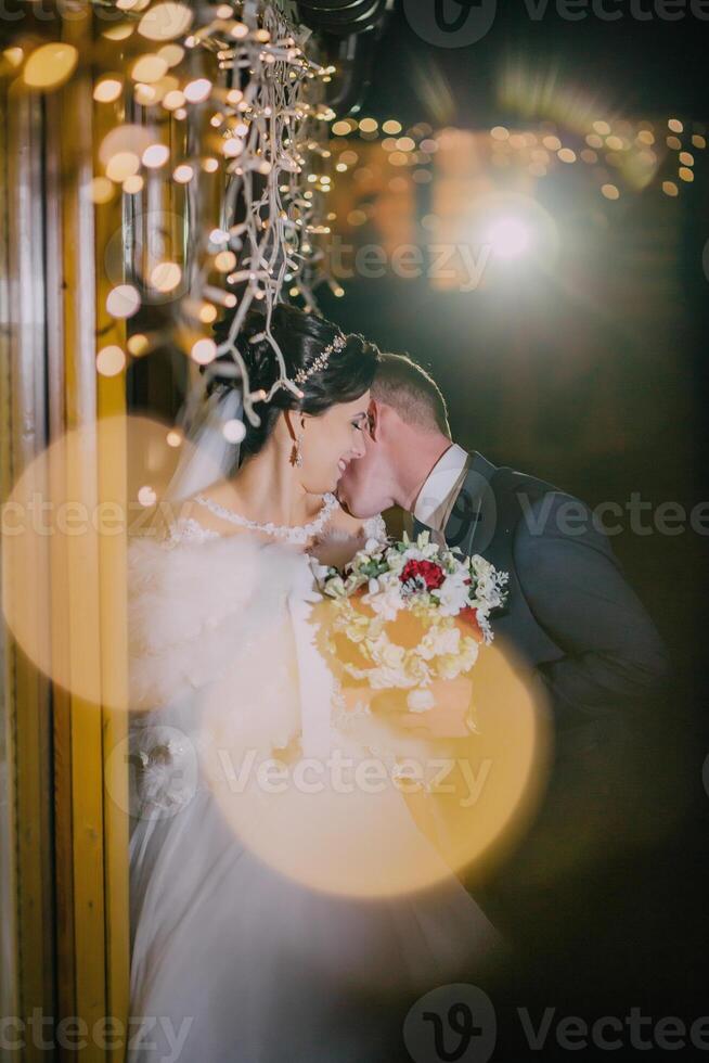 A bride and groom are kissing in front of a window with lights shining through photo