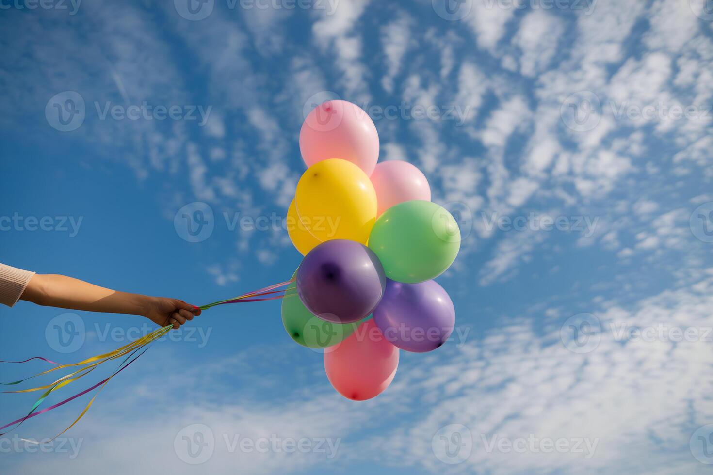 Arm holds colorful balloons against clear blue sky with scattered clouds photo