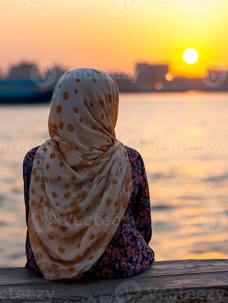 Contemplative Woman in Hijab Watching River Sunset with City Skyline photo